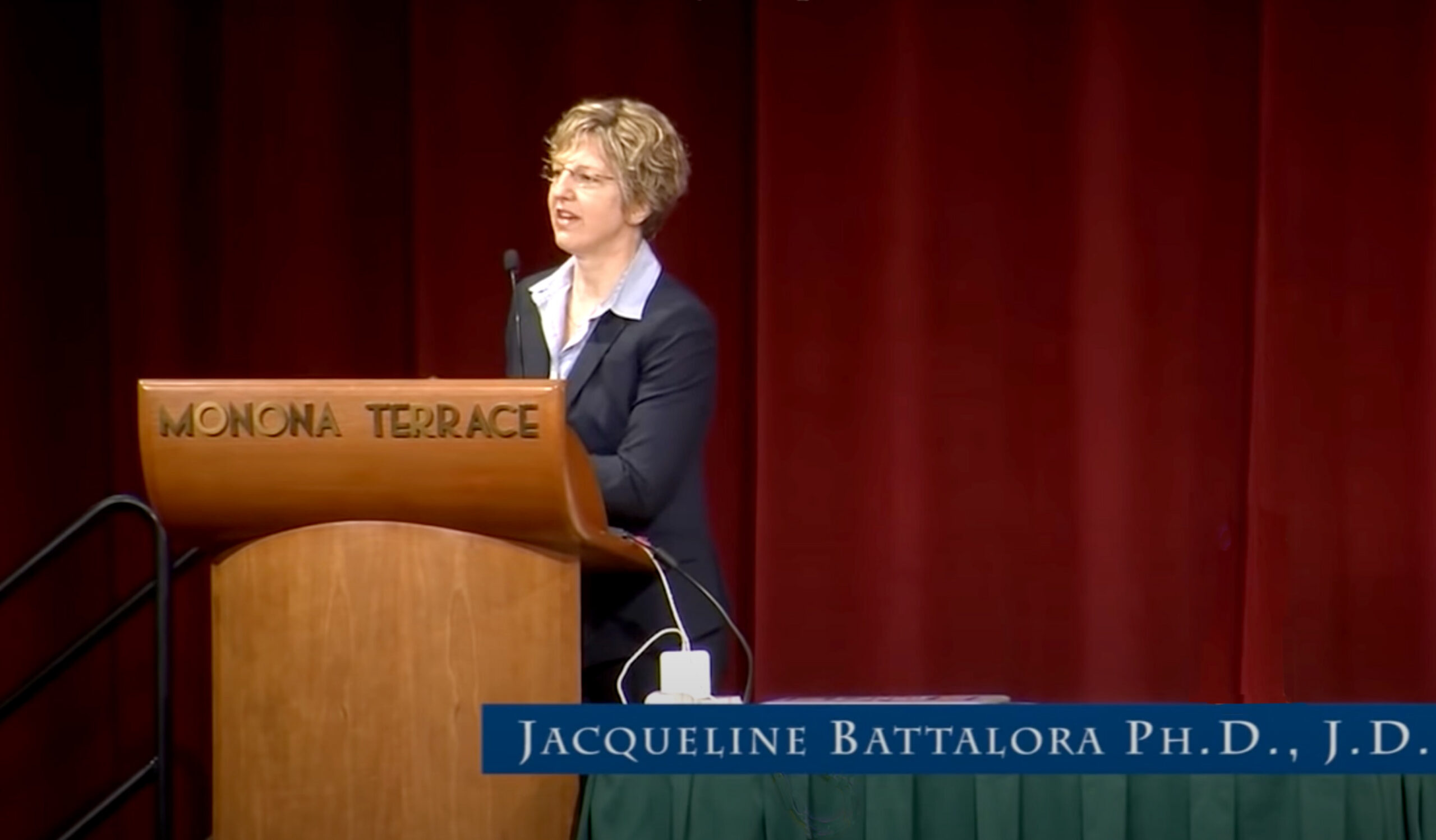 Photo captipned Jacqueline Battalora PH.D., J.D., white, blond haired woman with white button up shirt and grey suit speaking before a podium.